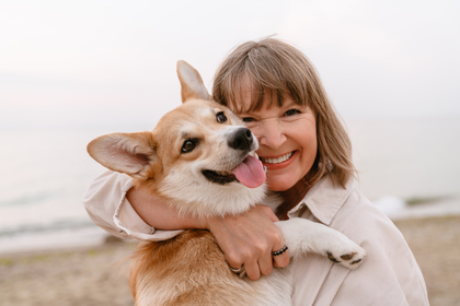 Kvinna kramar en glad corgi på stranden, båda ler mot kameran.