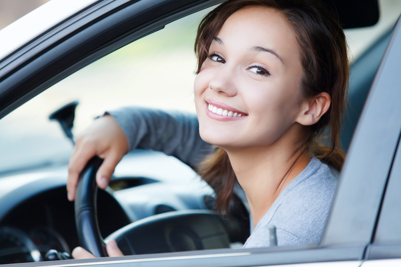 Ung kvinna sitter i en bil, håller i ratten och ler mot kameran.