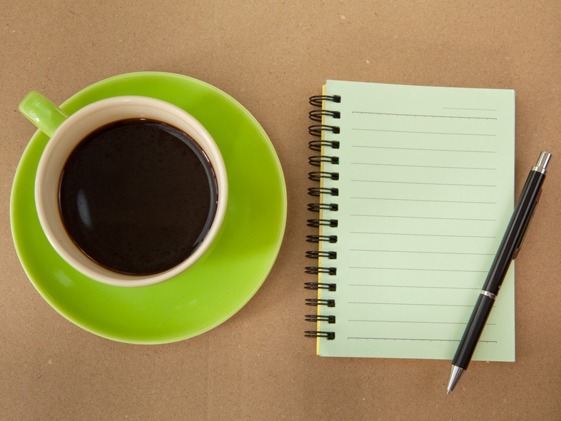 En kopp svart kaffe på ett grönt fat bredvid ett tomt spiralblock och en penna på ett brunt bord.