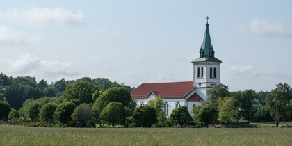 En vit kyrka med rött tak och grön spira står omgiven av träd på en äng under blå himmel.