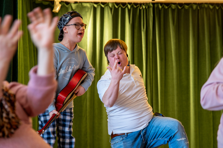 Två unga personer, en med gitarr, sjunger och gör rockposer framför gröna gardiner.