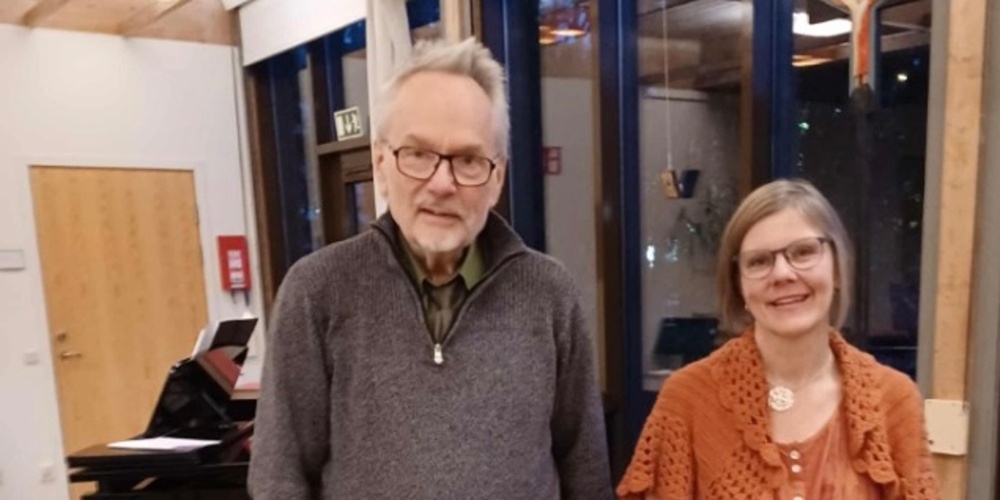 Två personer står inomhus bredvid ett piano, med stora fönster i bakgrunden.