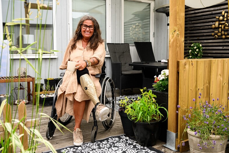 Kvinna med protesben sitter i rullstol på en uteplats omgiven av växter och trämöbler.