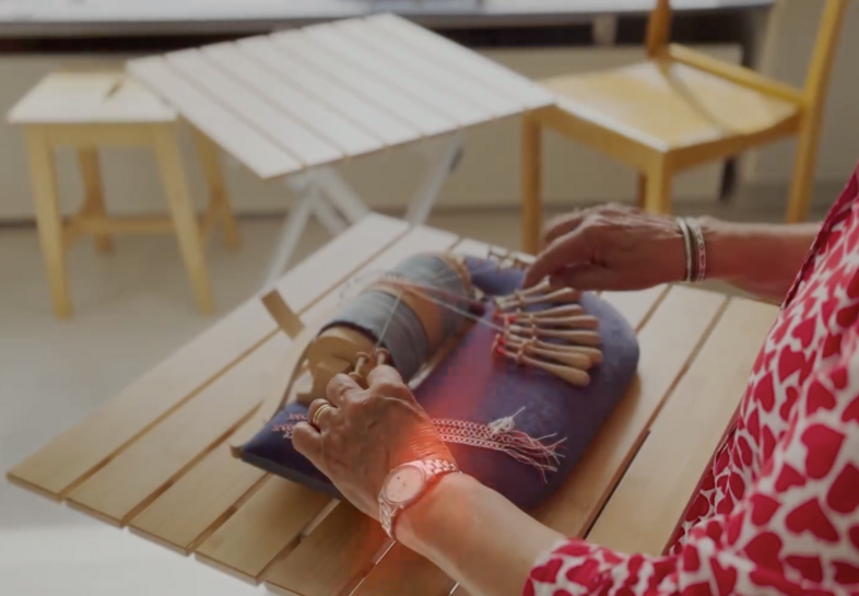 En person gör spetsknyppling på ett träbord, med flera stolar i bakgrunden.