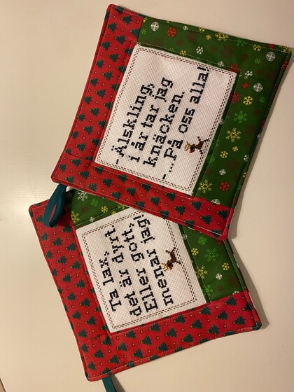 Två juliga grytlappar med broderade svenska texter och små renmotiv på ett bord.