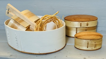 Tre runda askar i trä och björknäver står på ett bord, en av dem innehåller flätade band och träverktyg.