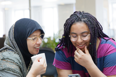Två unga kvinnor sitter tillsammans, ler och tittar på en mobiltelefon.