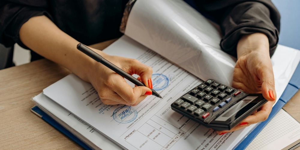 En person skriver på dokument med en penna och håller en miniräknare vid ett skrivbord.