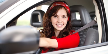 Ung kvinna med långt brunt hår och röd tröja sitter leende bakom ratten i en bil.