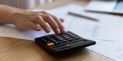 En person använder en miniräknare och arbetar vid en bärbar dator med papper på ett skrivbord.