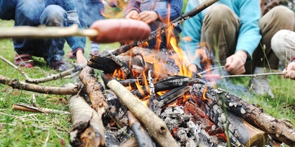 Flera personer grillar korv över öppen eld utomhus i en grön skogsmiljö.