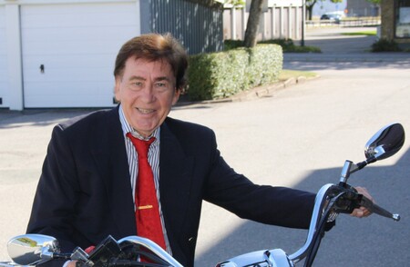Äldre man i kostym och röd slips sitter på en motorcykel utomhus vid en gata.