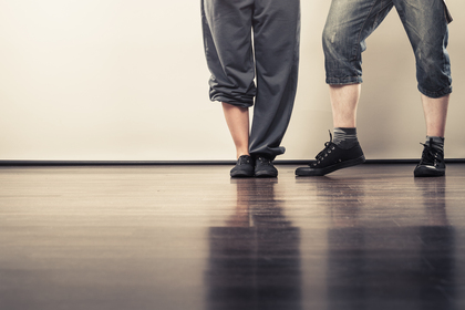 Två personer står på ett trägolv, endast deras ben och fötter syns. De bär avslappnade kläder och sneakers.