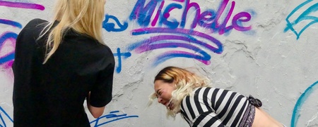 Två personer står framför Graffitiväggen i Tybbleparken. En av dem skrattar, den andra står med ryggen mot kameran. Väggen är vit, med text i rosa och lila, det står michelle. 