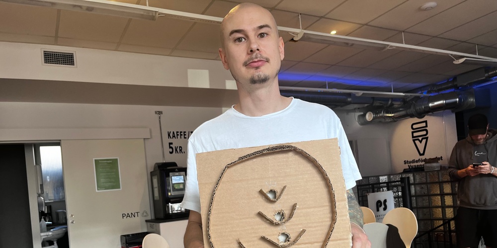 En person håller upp en kartong med konstverk av wellpapp i ett uppehållsrum med stolar och kaffemaskin.