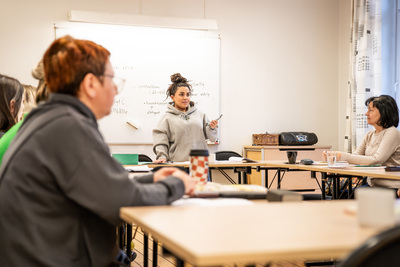En kvinna står vid en whiteboard och undervisar en grupp vuxna i ett klassrum.