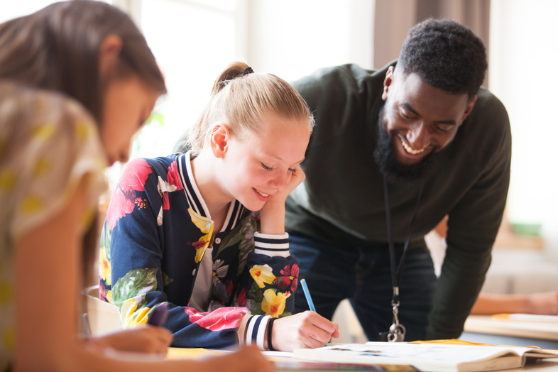 En lärare hjälper två elever med skolarbete vid ett bord i ett ljust klassrum.