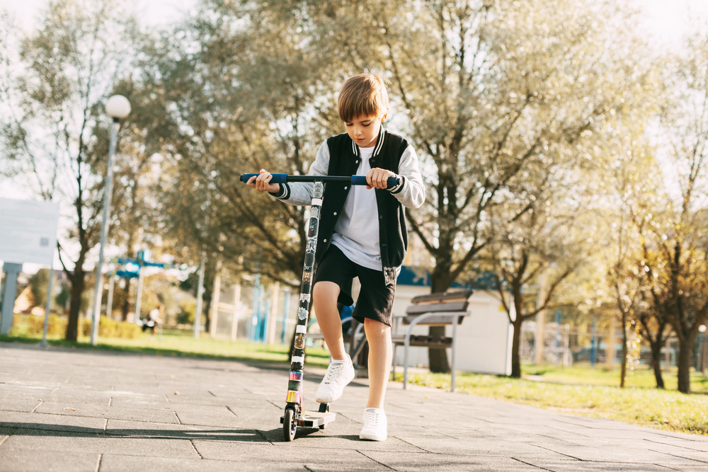 En pojke åker sparkcykel på en asfalterad gångväg i en park under en solig dag.