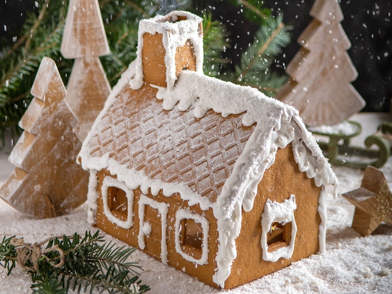 Ett pepparkakshus dekorerat med vit glasyr står i snöig miljö med granar och träd i bakgrunden.
