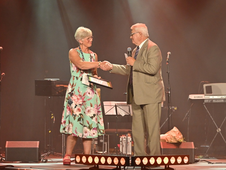 Ulrika Heie och Björn Körlof skakar hand på en scen under en prisutdelning, med mikrofoner och instrument i bakgrunden.