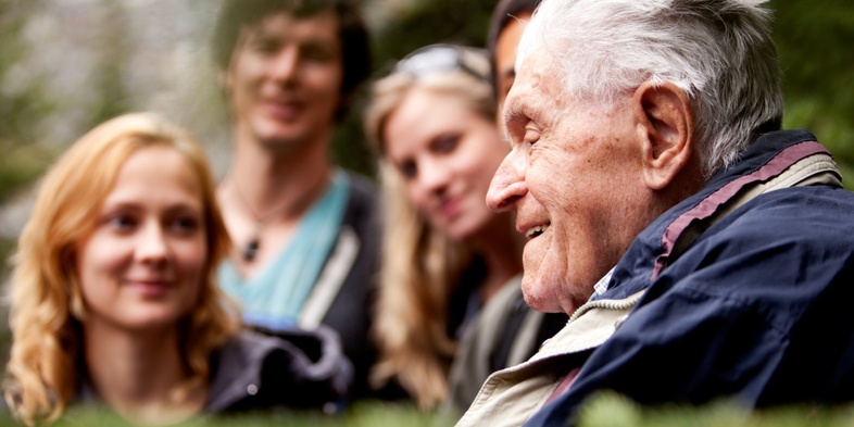 En äldre man pratar med en grupp yngre vuxna utomhus i en grönskande skogsmiljö.