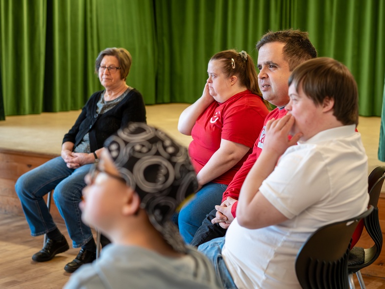 Fem personer sitter på stolar i en rad i ett rum med trägolv och grön scenbakgrund.