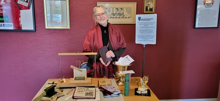 Kvinna med glasögon står bakom ett bord med böcker och föremål, mot en röd vägg med tavlor.