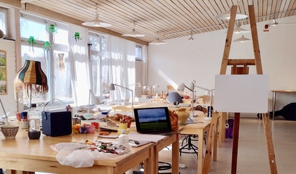 Ljust konstnärsateljé med arbetsbord, staffli, målarfärger och dator på borden.