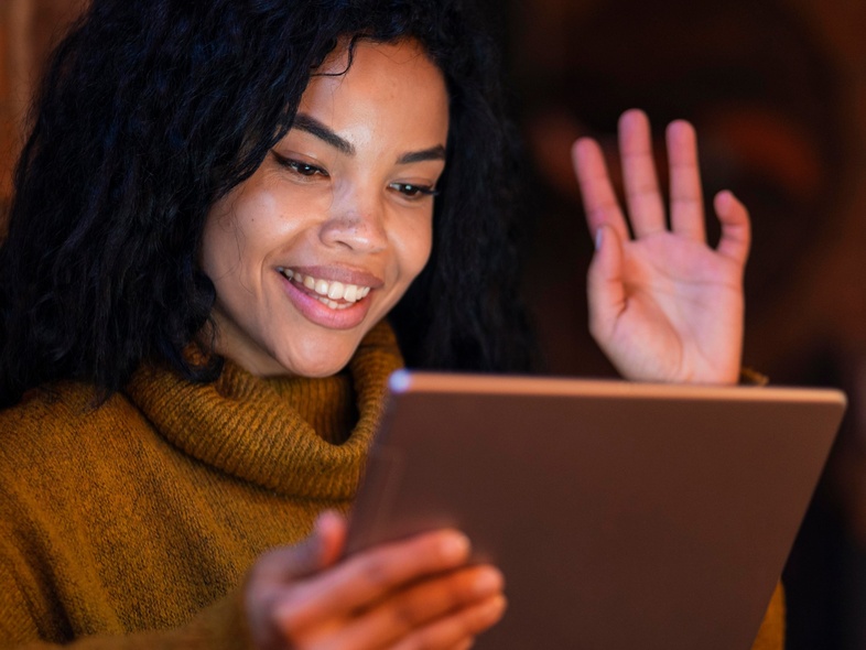 Kvinna med lockigt hår vinkar och ler mot en surfplatta i ett varmt upplyst rum.