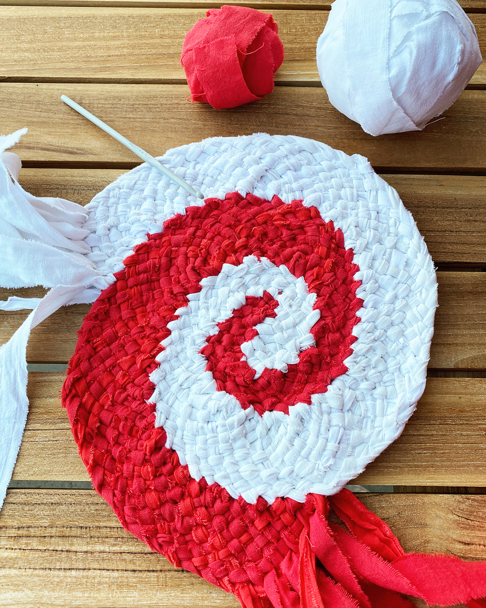 En virkad spiralmatta i rött och vitt ligger på ett träbord med garnbollar och en virknål bredvid.