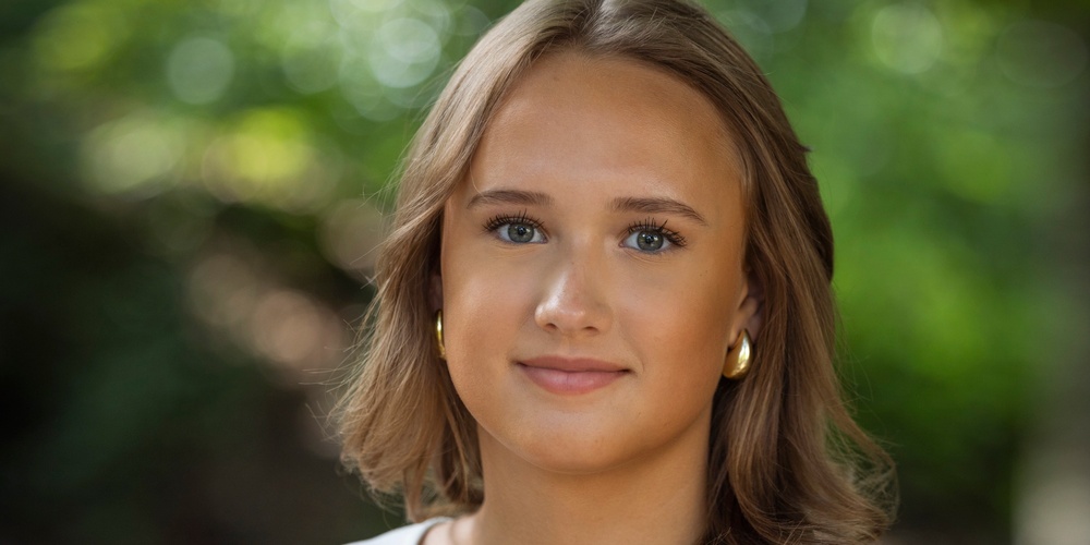 Person med axellångt brunt hår och vit topp står utomhus med grönskande bakgrund.