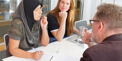 Tre personer sitter vid ett bord och diskuterar, med anteckningsblock och glas vatten framför sig.