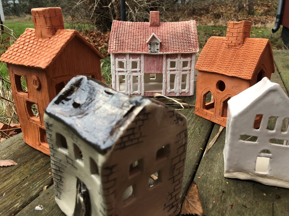 Fem små keramikhus står på ett träbord utomhus, omgivna av höstlöv och natur.