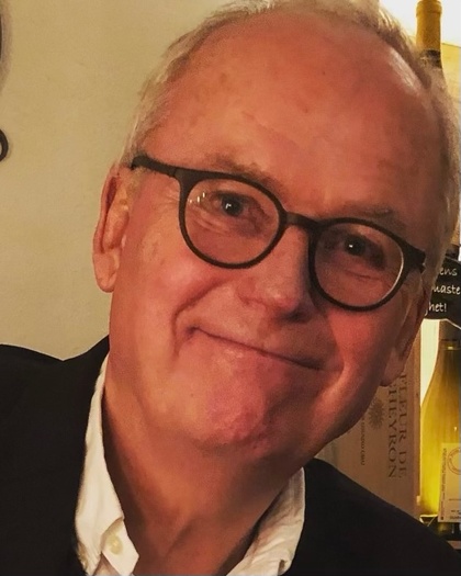 Äldre man med glasögon och vit skjorta ler mot kameran, inomhusmiljö med flaskor i bakgrunden.