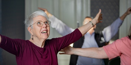 Äldre kvinna med glasögon sträcker ut armarna i ett rum, deltar i gruppaktivitet.