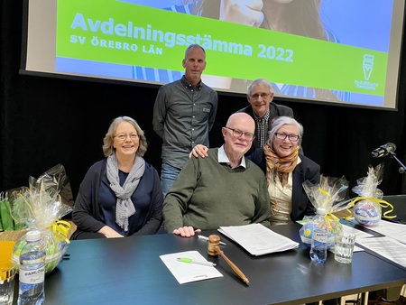 Fem personer sitter och står vid ett bord under ett möte, med en presentation i bakgrunden.