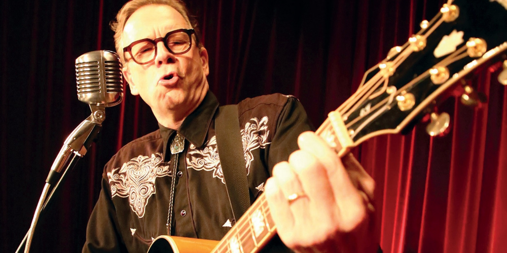 En person spelar gitarr och sjunger i mikrofon på scen med röda gardiner i bakgrunden.