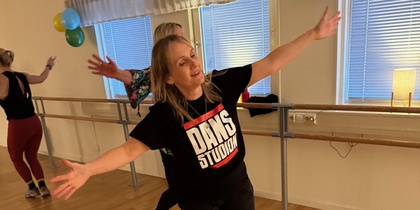 Kvinna dansar glatt i en dansstudio med trägolv och speglar, iförd svart t-shirt med texten 'DANS STUDIO'.