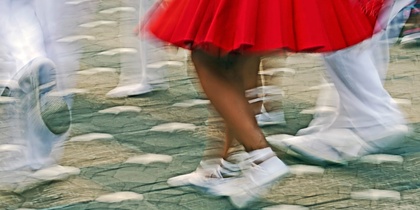 Personer dansar i rörelse, klädda i vita kläder och röd kjol, på en stenlagd gata.