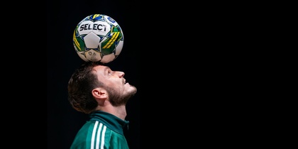 En man i grön sportjacka balanserar en fotboll på pannan mot svart bakgrund.
