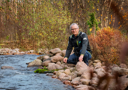 En man sitter på stenar vid en bäck i en höstig skogsmiljö.