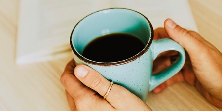 En person håller en blå mugg med kaffe framför en öppen bok på ett bord med pumpor i bakgrunden.