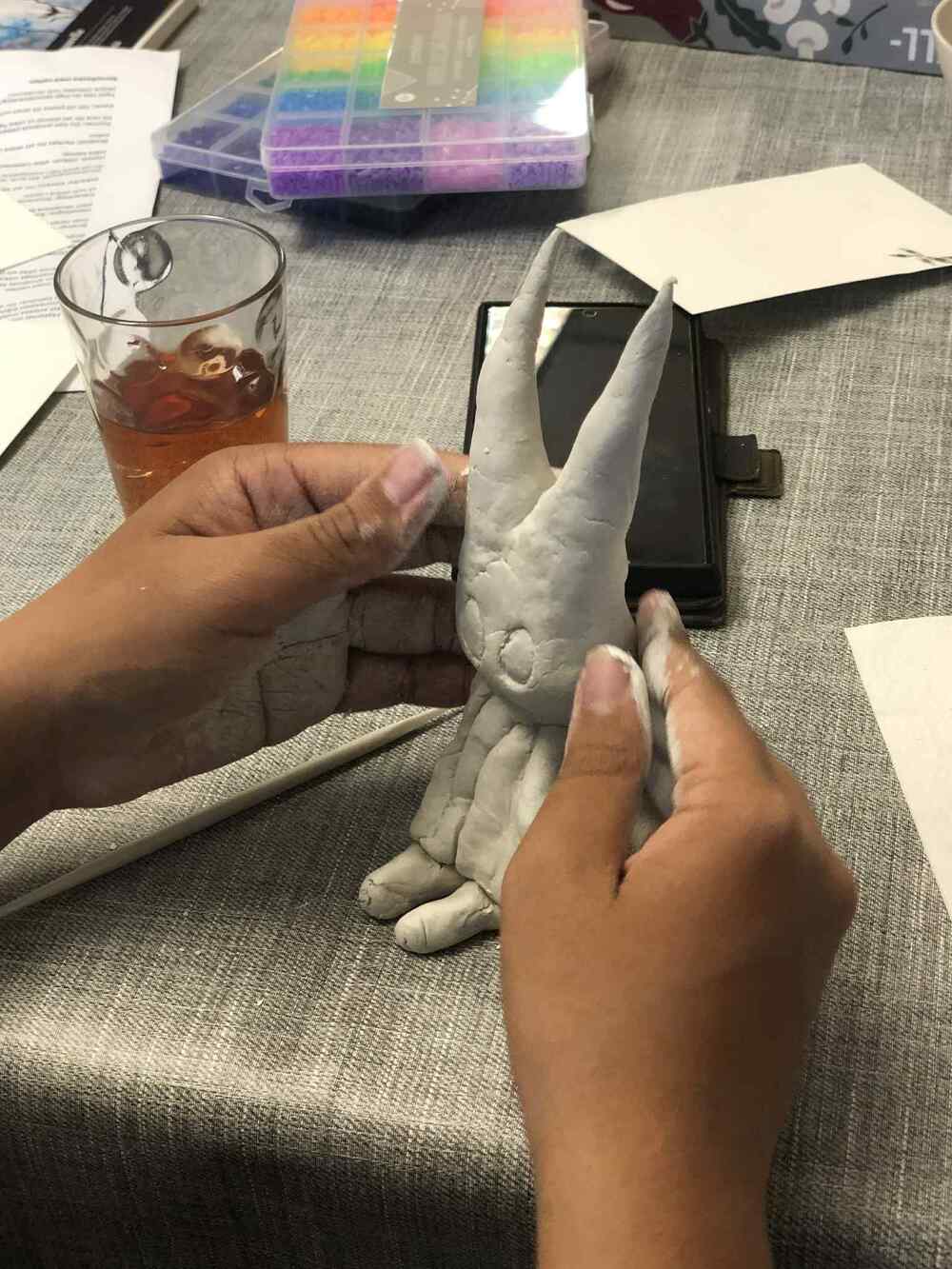 Två händer formar en kaninliknande figur av lera på ett bord med verktyg och ett glas dryck.