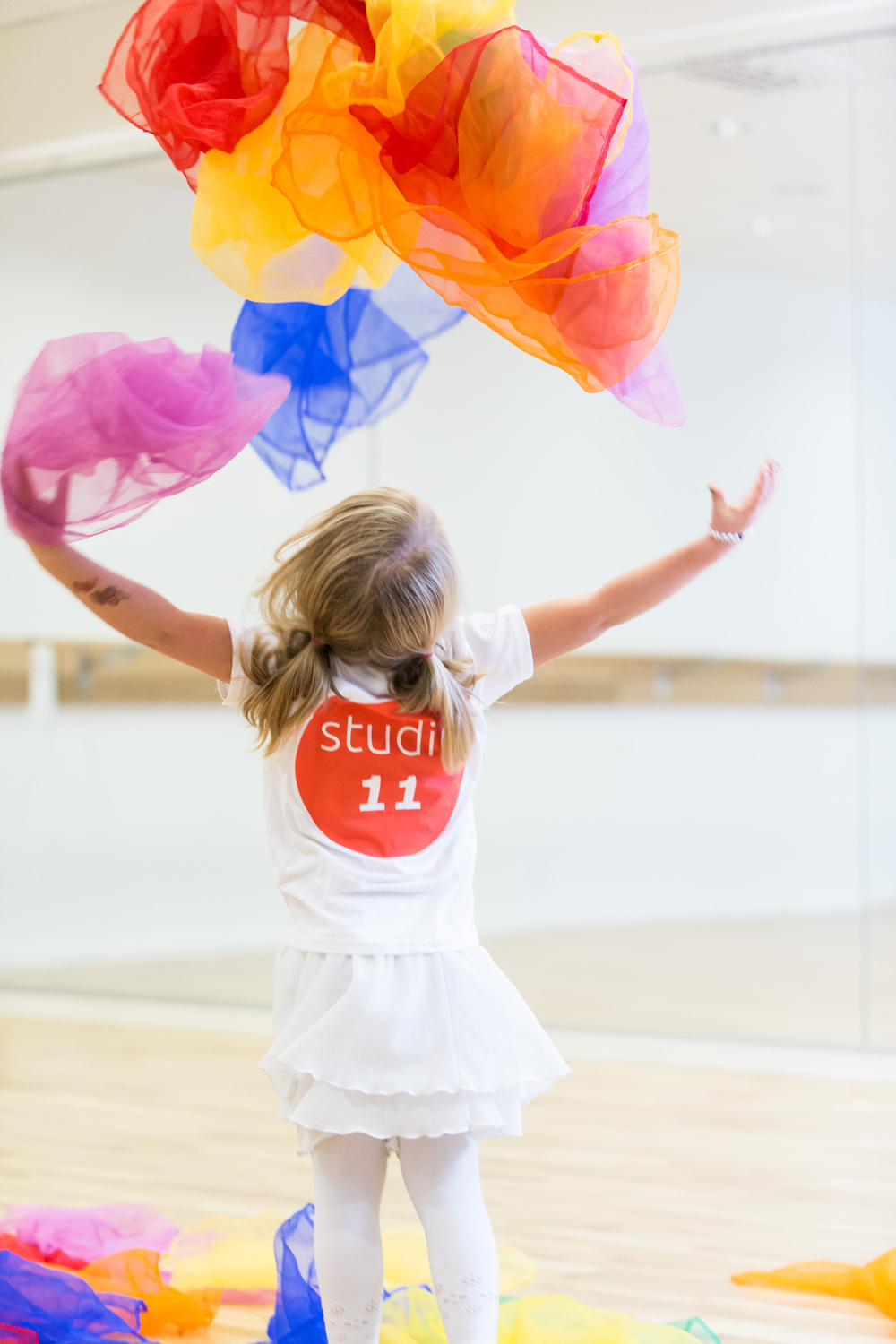 Ett barn i vit klänning kastar färgglada tygsjalar i luften i en ljus dansstudio.