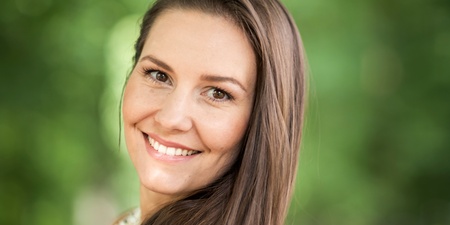 Leende kvinna med långt brunt hår står utomhus mot en suddig grön bakgrund.