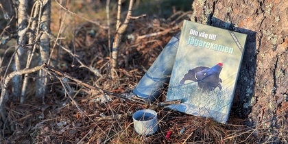 En bok med en fågel på omslaget, en termos och en kopp står lutade mot ett träd i skogen.