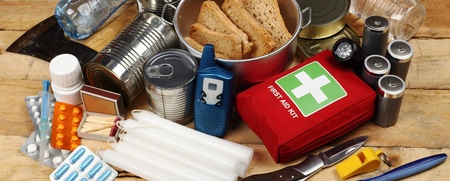 Nödutrustning med konserver, vattenflaskor, första hjälpen-kit, ficklampa, kniv, ljus och mediciner på ett träbord.