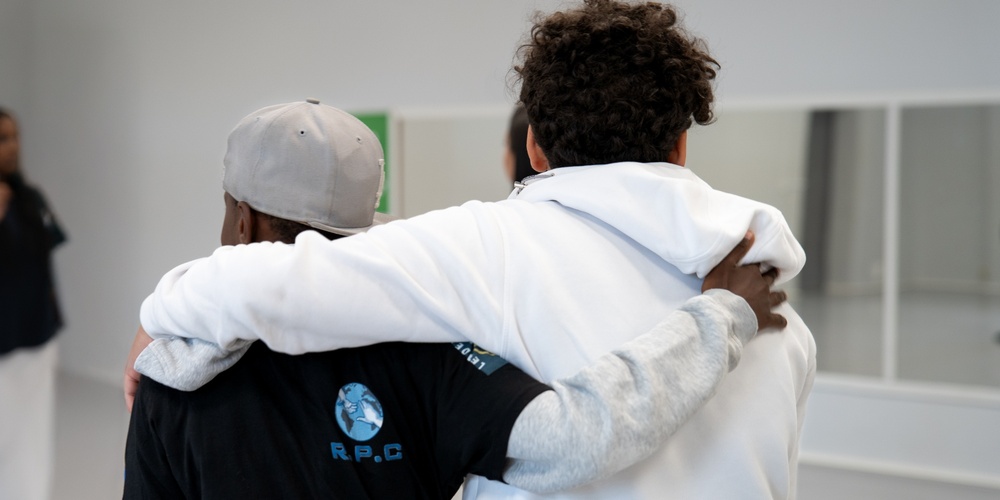 Två personer står med armarna om varandra i ett ljust rum, en tredje person syns i bakgrunden.