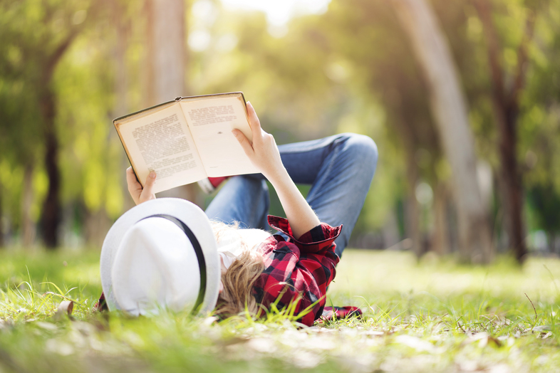 Person med hatt ligger på gräset utomhus och läser en bok i solskenet.