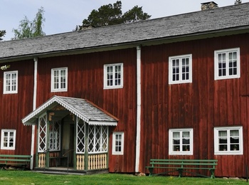 Fågelsjö gammelgård bortom Åa en röd Hälsingegård med vita fönster och veranda, omgiven av gräs och träd under en ljus himmel.